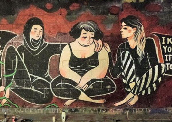 Graffito mit 3 Frauen, die nebeneinander auf dem Boden sitzen. Die beiden außen sitzenden Personen legen jeweils eine Hand auf die Person in der Mitte. Die beiden äußeren Personen sagen: "That's not ok and it's not your fault" und "I know how you feel it happened to me too".