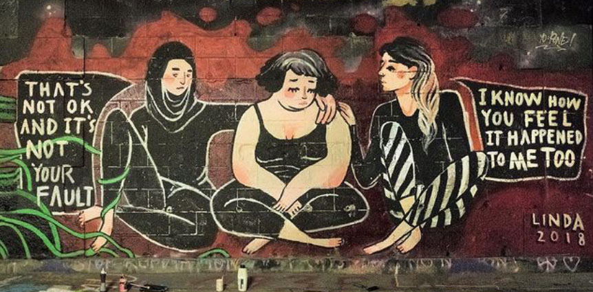 Graffito mit 3 Frauen, die nebeneinander auf dem Boden sitzen. Die beiden außen sitzenden Personen legen jeweils eine Hand auf die Person in der Mitte. Die beiden äußeren Personen sagen: "That's not ok and it's not your fault" und "I know how you feel it happened to me too".