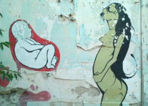 Gemälde auf einer alten Hauswand. Rechts ist eine Person mit langen Haaren, die eine Hand auf ihren Bauch legt, auf der linken Seite ist ein Embryo, der sich in einer roten Blase befindet.