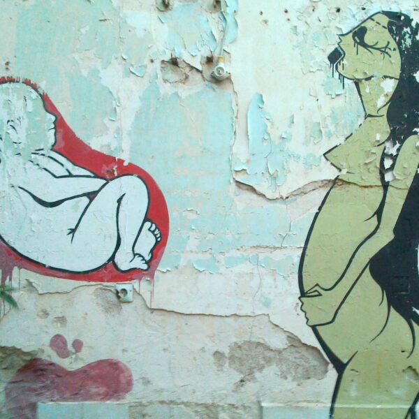 Gemälde auf einer alten Hauswand. Rechts ist eine Person mit langen Haaren, die eine Hand auf ihren Bauch legt, auf der linken Seite ist ein Embryo, der sich in einer roten Blase befindet.