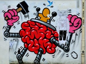 Graffito eines roten Hirns mit Beinen und Armen und einer orangen Figur, die im Gehirn sitzt und zwei Schalthebel bedient. Das rote Hirn hat an den Armen pinke Hände, eine Hand ist zur Faust geballt, die andere hält einen Hammer.