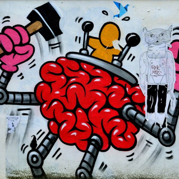 Graffito eines roten Hirns mit Beinen und Armen und einer orangen Figur, die im Gehirn sitzt und zwei Schalthebel bedient. Das rote Hirn hat an den Armen pinke Hände, eine Hand ist zur Faust geballt, die andere hält einen Hammer.