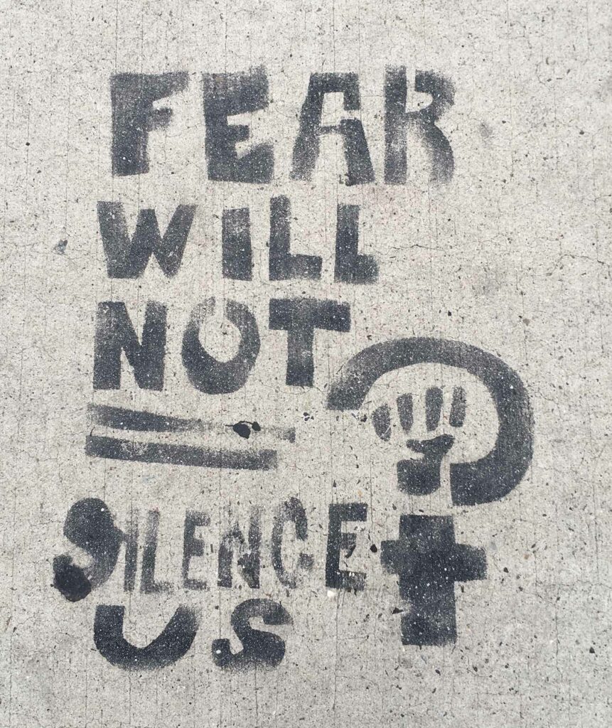 Schablonen-Graffito am Boden mit dem Schriftzug "Fear will not silence us" und einem Frauenzeichen mit geballter Faust.