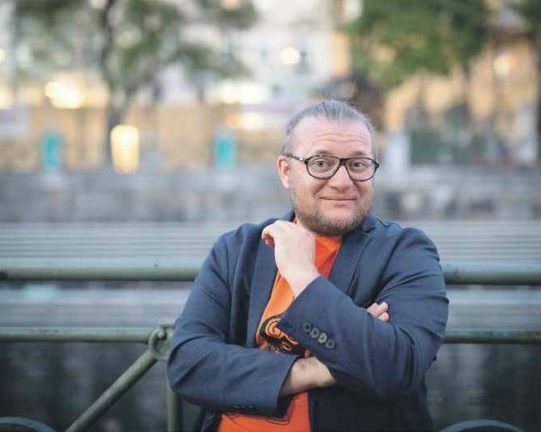 Foto von Persson Baumgartinger. Baumgartinger trägt eine Brille, ein graues Sakko und ein oranges T-Shirt.