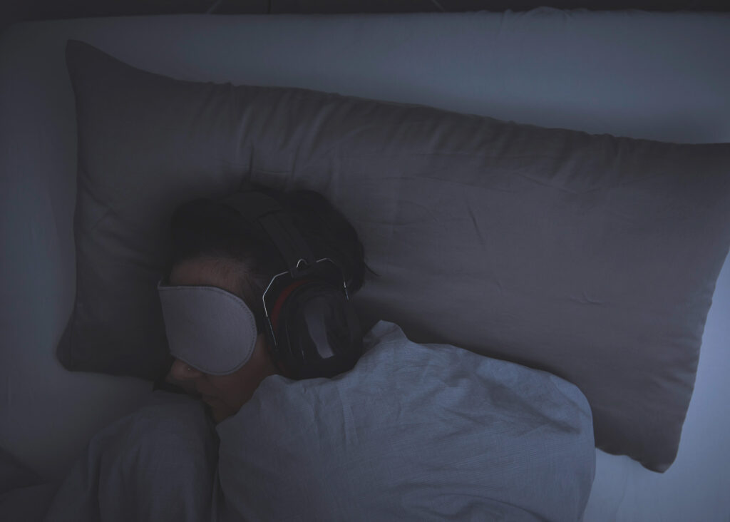 Eine Person mit Augenbinde und Kopfhörern liegt in einem Bett in einem dunklen Zimmer.