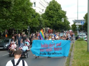 Menschenmenge bei der Pride Parade Berlin, im Vordergrund Rollstuhlfahrer*innen, eine Regenbogenfahne und ein großes Transparent mit dem Text "behindert und verrückt feiern Pride Parade Berlin"