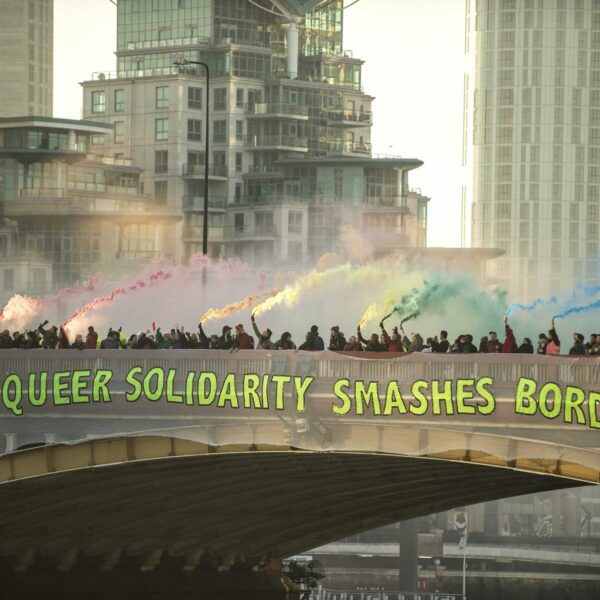 Menschenmenge auf einer Brücke mit Rauchfackeln in Regenbogenfarben, auf der Brücke hängt ein großes Transparent mit dem Text "Queer Solidarity Smashes Borders"