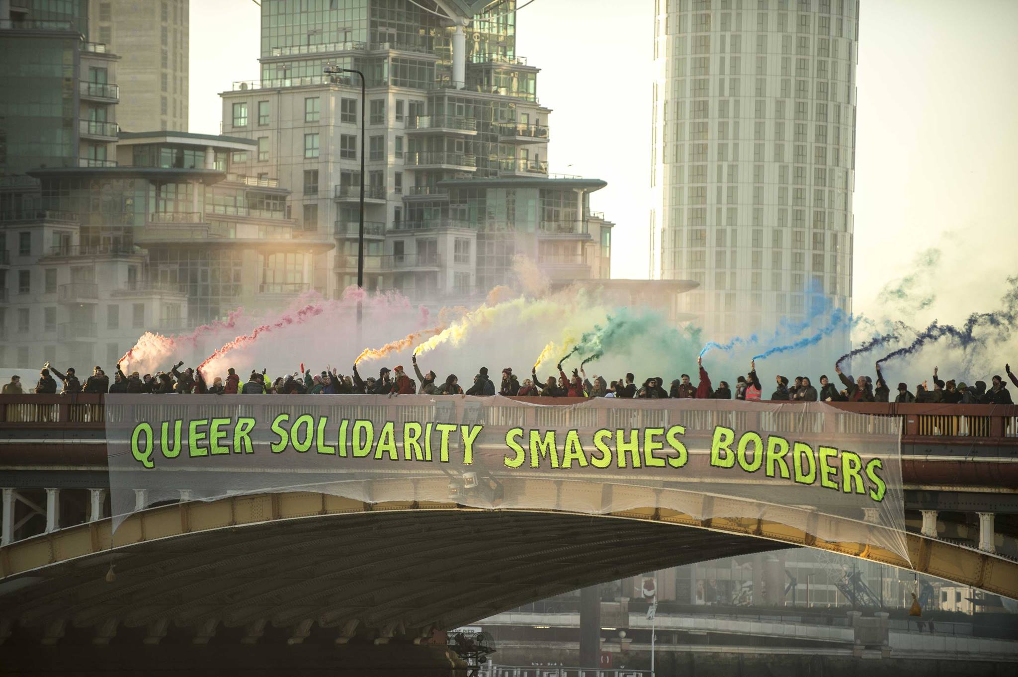 Menschenmenge auf einer Brücke mit Rauchfackeln in Regenbogenfarben, auf der Brücke hängt ein großes Transparent mit dem Text 