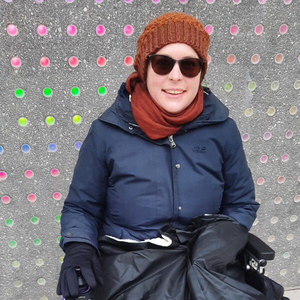 Sarah Kanawin sitzt in einem Elektrorollstuhl. Sie lächelt und trägt eine Sonnenbrille, braune Haube und Schal, dunkelblaue Jacke, schwarze Handschuhe und hat eine Decke über den Beinen. Im Hintergrund eine Wand mit vielen bunten Punkten.