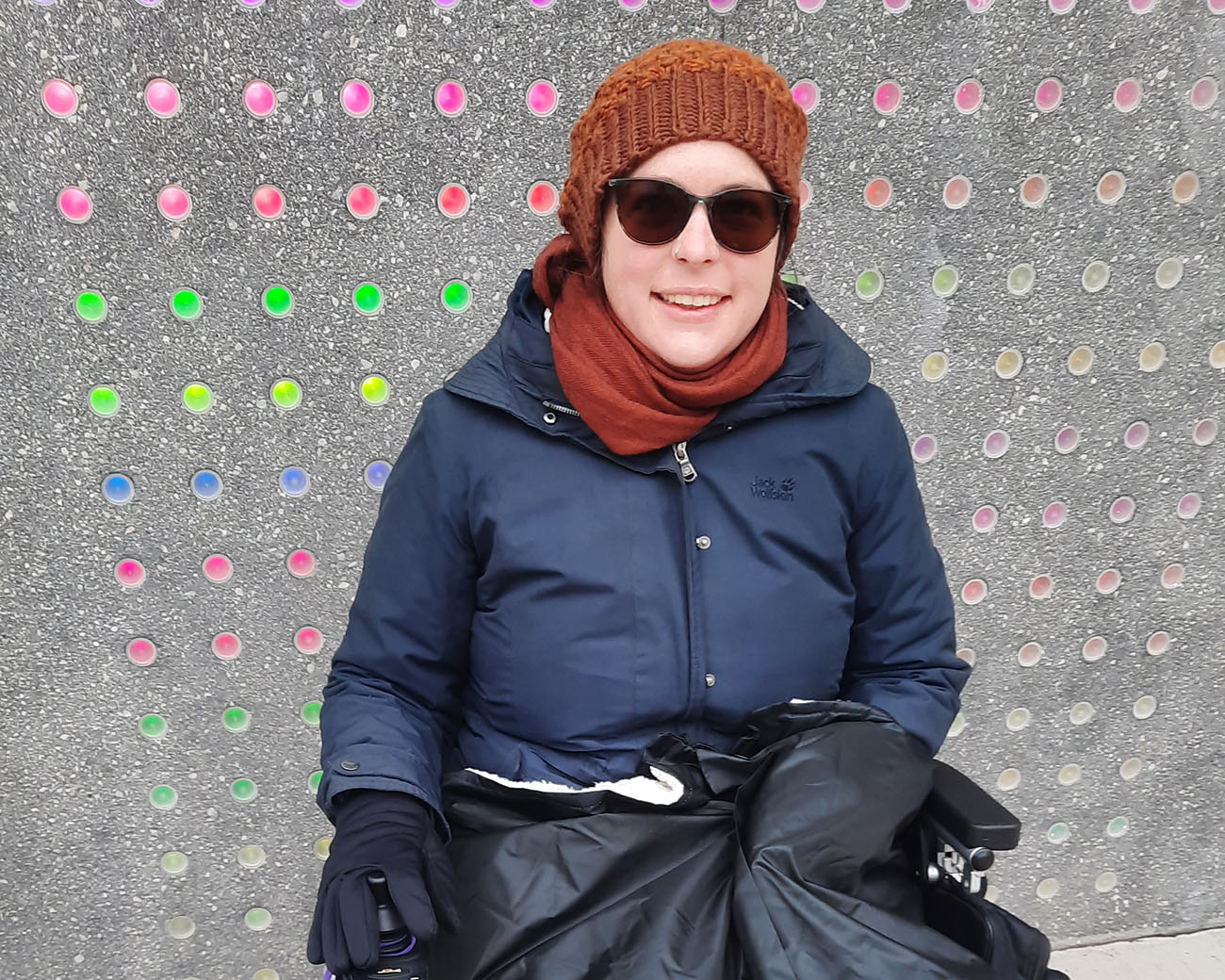 Sarah Kanawin sitzt in einem Elektrorollstuhl. Sie lächelt und trägt eine Sonnenbrille, braune Haube und Schal, dunkelblaue Jacke, schwarze Handschuhe und hat eine Decke über den Beinen. Im Hintergrund eine Wand mit vielen bunten Punkten.