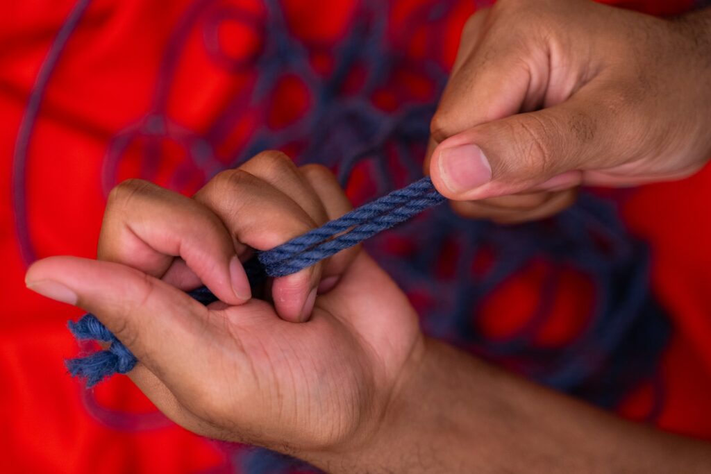blaues Seil, das zwischen zwei Händen gespannt ist, der Hintergrund ist verschwommen rot mit in Schlingen liegendem blauem Seil