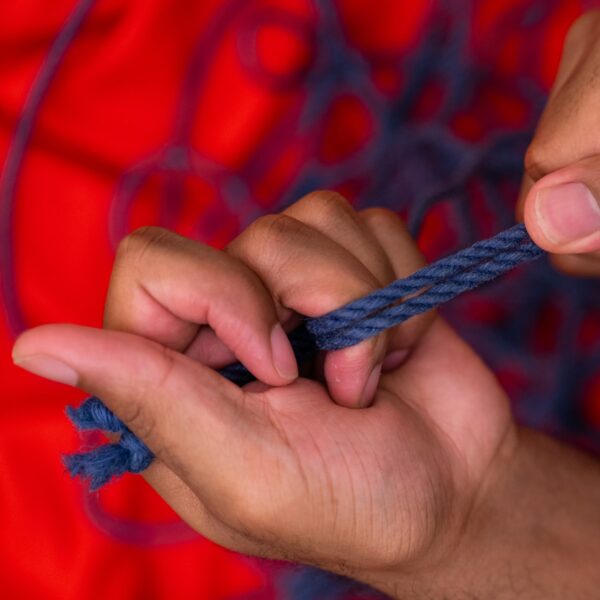 blaues Seil, das zwischen zwei Händen gespannt ist, der Hintergrund ist verschwommen rot mit in Schlingen liegendem blauem Seil