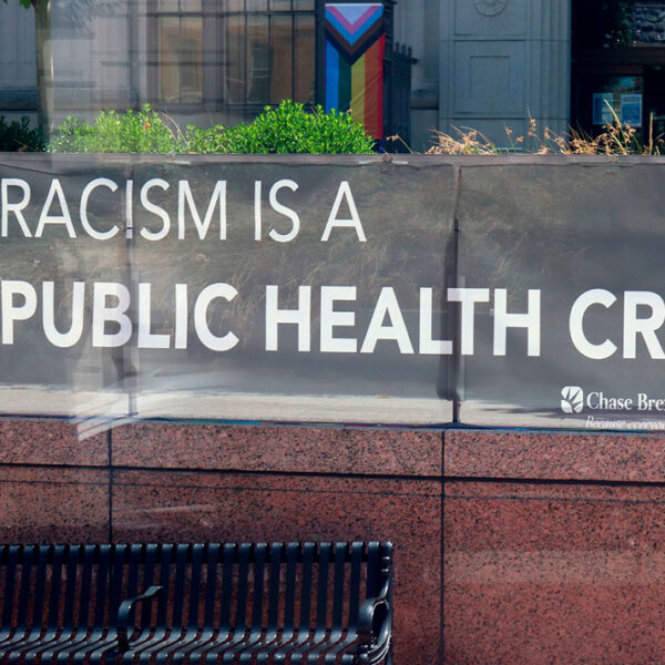Banner im öffentlichen Raum mit dem Text: Racism is a public health crises und dem Logo von Chase Brexton Health Care. Im Hintergrund ist eine Progress-Pride-Flagge sichtbar.