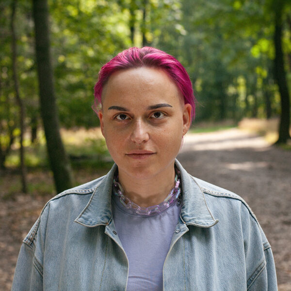 Porträtfoto von Silence Conrad im Wald. Silence hat pinke, nach hinten gekämmte Haare, ein Septumpiercing, trägt eine offene Jeansjacke mit lila T-Shirt und lila Kette um den Hals und blickt in die Kamera.