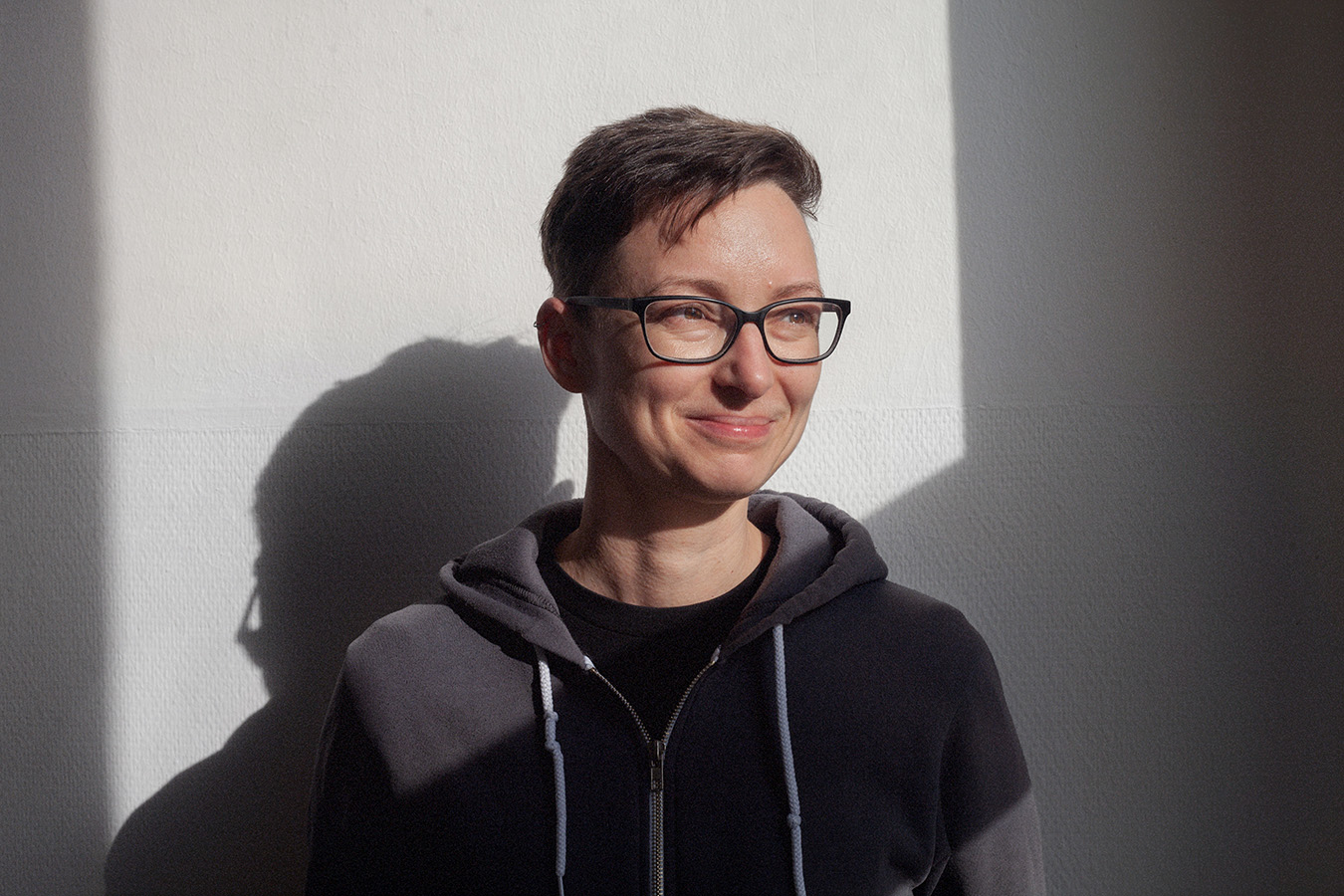 Porträtbild von Bettina Enzenhofer. Bettina hat kurze braune Haare, trägt eine Brille und einen grauen Kaputzenpullover und lächelt