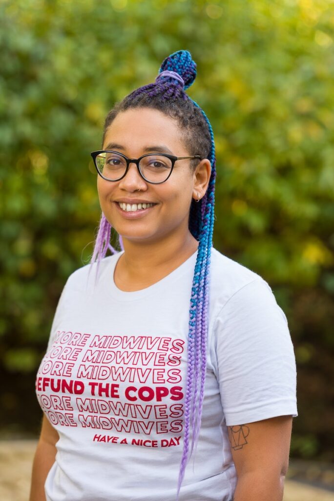 Foto von Laura Warria. Laura trägt ein T-Shirt mit der Aufschrift "Defund the cops. More midwifes. Have a nice day", eine Brille mit dunklem Rahmen und hat Rastazöpfe in dunkelbraun, türkis und lila. Laura ist Schwarz und lächelt in die Kamera.