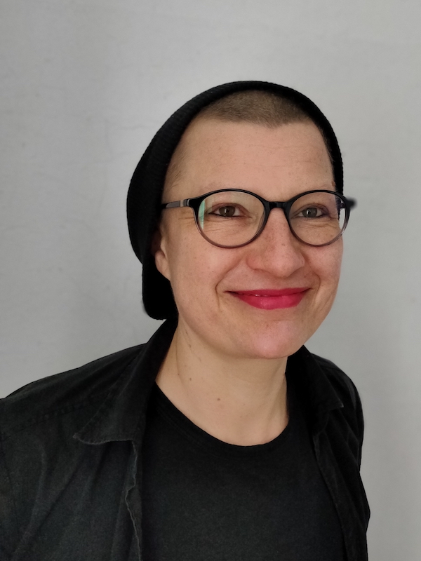 Porträtfoto von Kirsten Achtelik. Kirsten Achtelik trägt ein schwarzes Oberteil, eine schwarze Haube über kurz rasierten Haaren, eine Brille mit dunklem Rahmen und roten Lippenstift. Kirsten Achtelik lächelt in die Kamera.