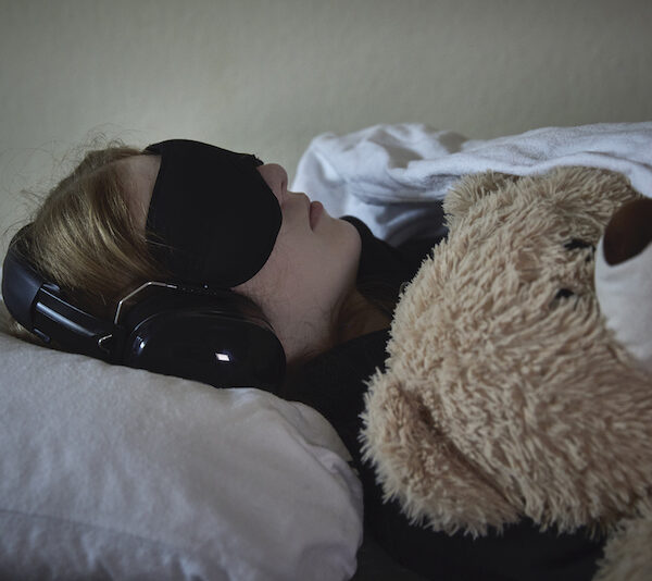 Ein Kind liegt im Bett, es trägt eine Schlafmaske und Kopfhörer. Daneben liegt ein Teddy.