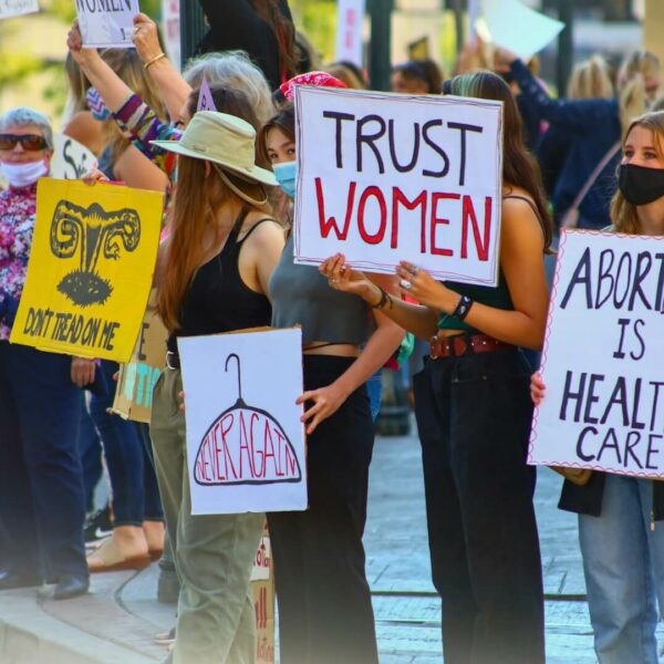 Mehrere Personen mit Demoschildern in der Hand, auf den Schildern steht unter anderem der Text "abortion is health care" und "trust women", auf einem Schild ist ein Kleiderbügel gezeichnet und der Text "never again" geschrieben.