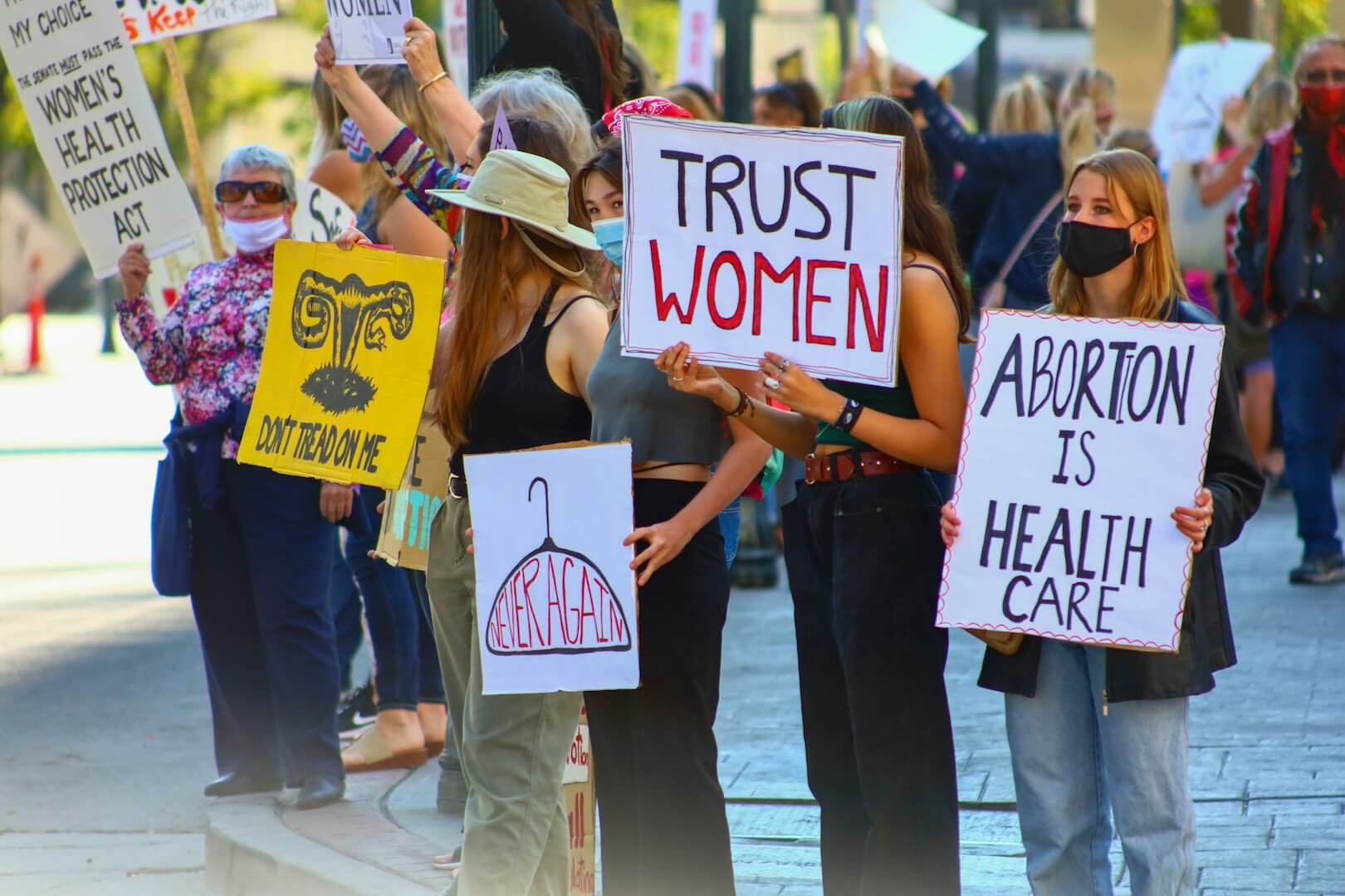 Mehrere Personen mit Demoschildern in der Hand, auf den Schildern steht unter anderem der Text "abortion is health care" und "trust women", auf einem Schild ist ein Kleiderbügel gezeichnet und der Text "never again" geschrieben.