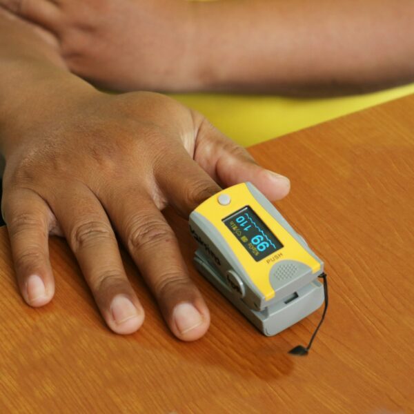 Eine Hand mit dunkler Hautfarbe liegt auf einem Tisch, ein Pulsoxymeter misst an einem Finger den Sauerstoffgehalt.
