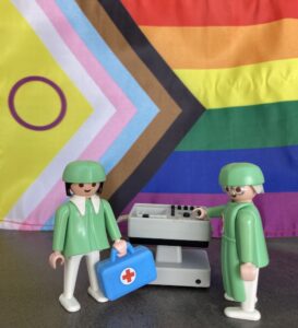 2 Legofiguren, die als Gesundheitspersonal gekleidet sind, stehen vor einer Regenbogenfahne