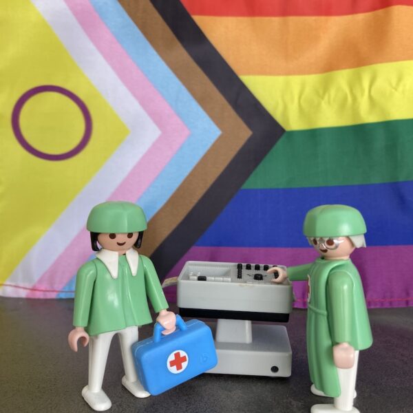 2 Legofiguren, die als Gesundheitspersonal gekleidet sind, stehen vor einer Regenbogenfahne