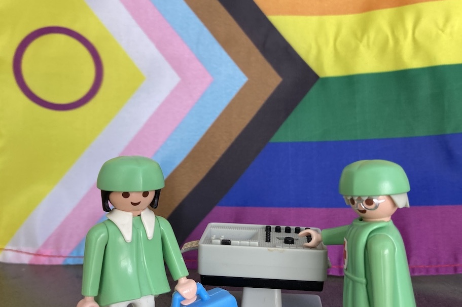 2 Legofiguren, die als Gesundheitspersonal gekleidet sind, stehen vor einer Regenbogenfahne