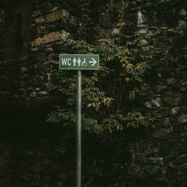 Ein WC-Schild im öffentlichen Raum mit einem Pfeil und Piktogrammen für Mann, Frau und Barrierefreiheit