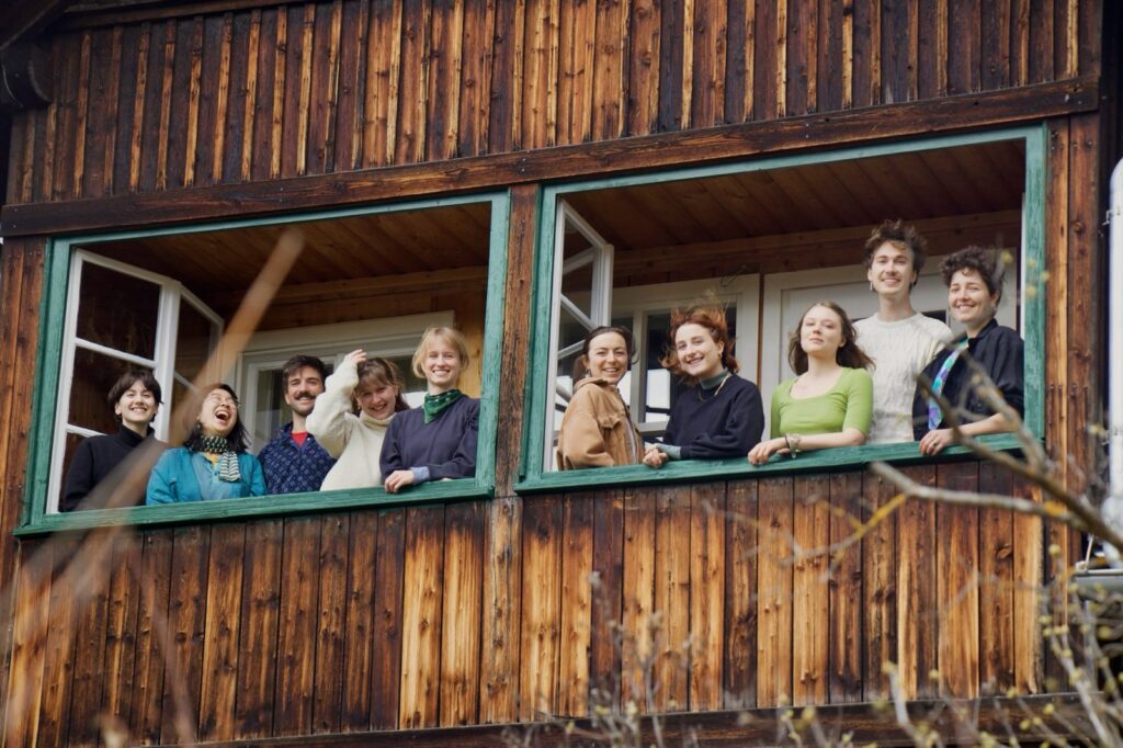 Foto mit 10 Personen, die in die Kamera lachen. Sie befinden sich in einem Holzhaus, vor geöffneten Fenstern und schauen nach Außen in die Kamera.