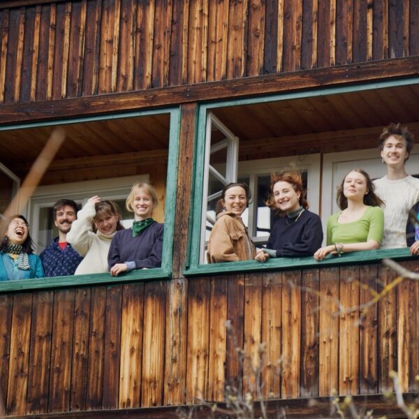 Foto mit 10 Personen, die in die Kamera lachen. Sie befinden sich in einem Holzhaus, vor geöffneten Fenstern und schauen nach Außen in die Kamera.