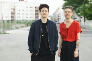 Foto von Ulrike Friedl und Yvonno Leeb von Tamo Bestattungen. Beide haben kurze Haare und eine Brille. Sie stehen auf einer Straße, im Hintergrund Wohnhäuser.