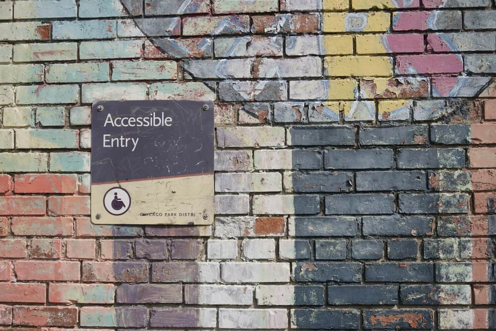 Mauer aus Ziegelsteinen mit Graffiti und einem Schild mit dem Text "Accessible Entry" und einem Piktogramm von einer Person im Rollstuhl