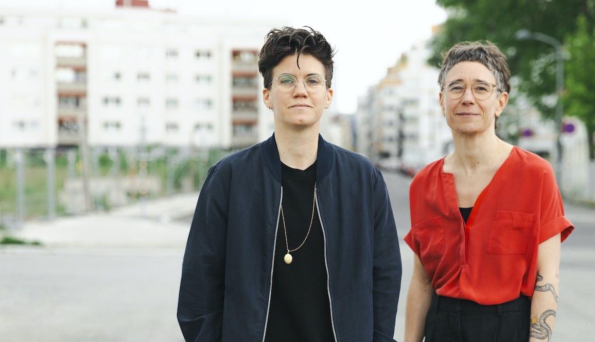 Foto von Ulrike Friedl und Yvonno Leeb von Tamo Bestattungen. Beide haben kurze Haare und eine Brille. Sie stehen auf einer Straße, im Hintergrund Wohnhäuser.