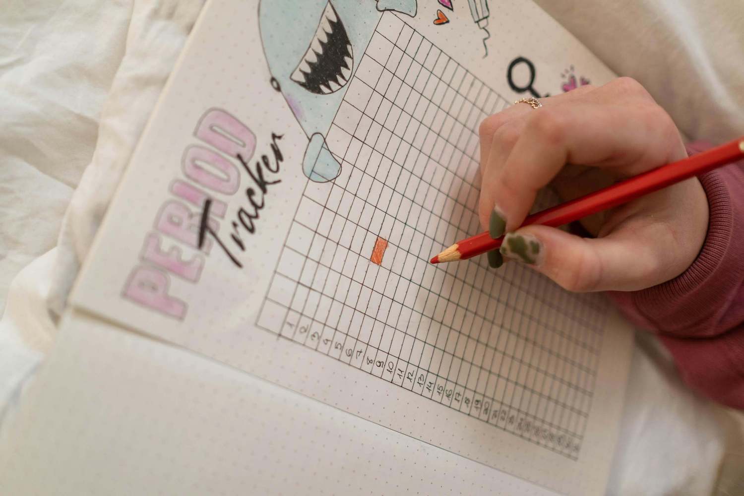 Blatt Papier mit der Überschrift "Period Tracker". Eingezeichnet sind 31 nummerierte Zeilen. Eine Hand hält einen roten Stift. Ein Feld auf dem Blatt ist rot ausgemalt.