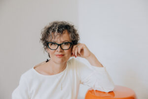Porträtfoto von Martina Eberhart. Sie hat kurze, graue, gelockte Haare, eine schwarz-türkise Brille, trägt ein weißes Oberteil und blickt in die Kamera.