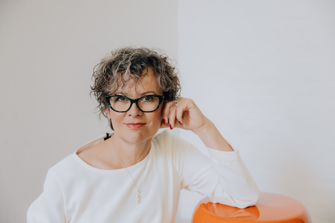 Porträtfoto von Martina Eberhart. Sie hat kurze, graue, gelockte Haare, eine schwarz-türkise Brille, trägt ein weißes Oberteil und blickt in die Kamera.