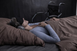 Eine Frau liegt im Bett, sie trägt eine Augenbinde und Gehörschutz. Neben dem Bett ist ein Rollstuhl.