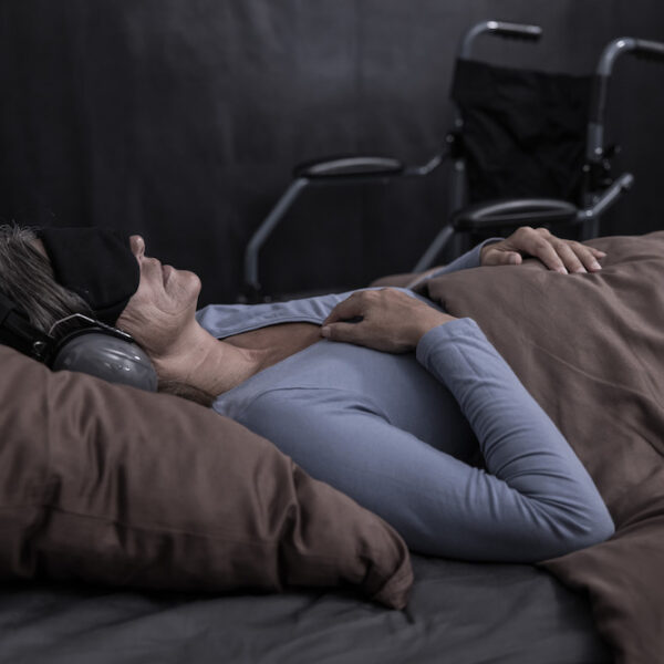 Eine Frau liegt im Bett, sie trägt eine Augenbinde und Gehörschutz. Neben dem Bett ist ein Rollstuhl.