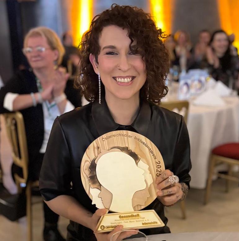 Patricia Amrita Pöchhacker hält die Skulptur des Österreichischen Inklusionspreises 2025 und lacht in die Kamera.
