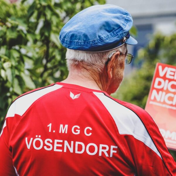 Eine Person von hinten fotografiert. Sie hat kurze graue Haare, trägt eine Brille und ein Hörgerät und hält in der Hand eine Broschüre mit dem Text "Vergiss dich nicht.".