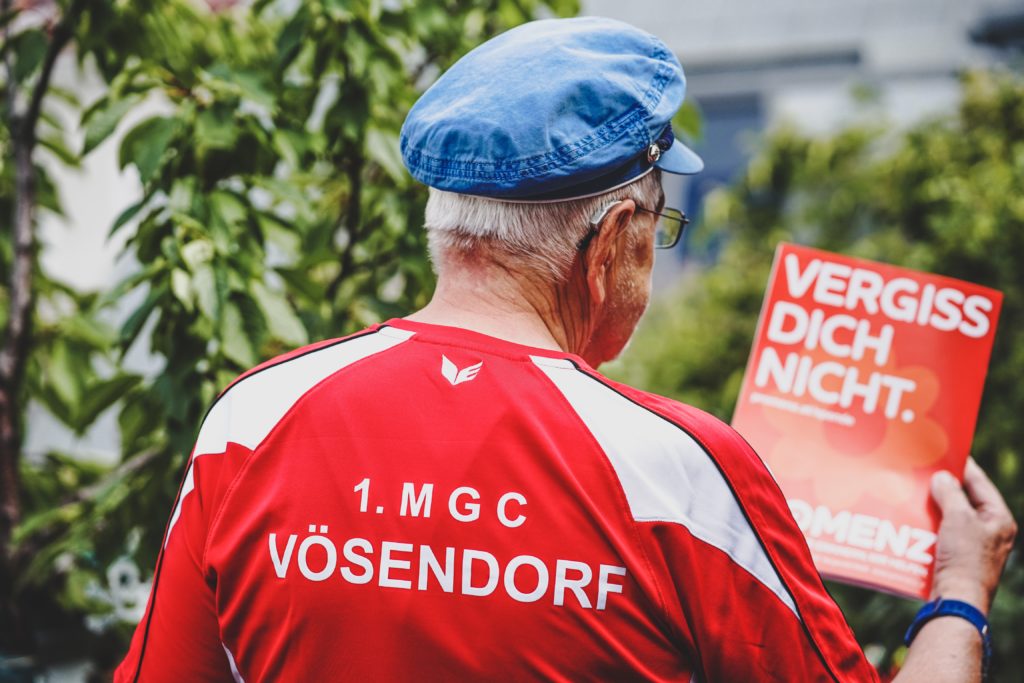 Eine Person von hinten fotografiert. Sie hat kurze graue Haare, trägt eine Brille und ein Hörgerät und hält in der Hand eine Broschüre mit dem Text "Vergiss dich nicht.".